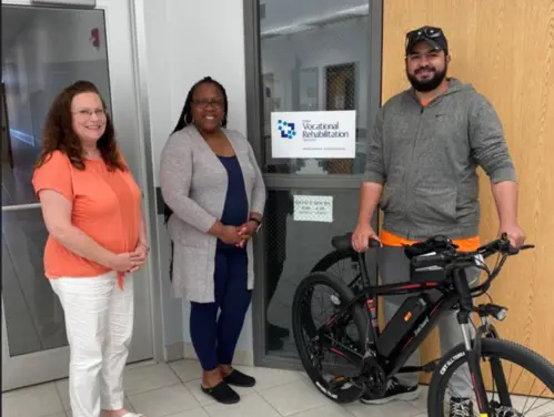 Three individuals standing next to each other and smiling. Person on the right is Jeremie Torres. He is standing behind a bike.