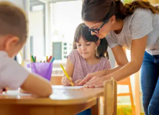 teacher bends over table to help child in classroom